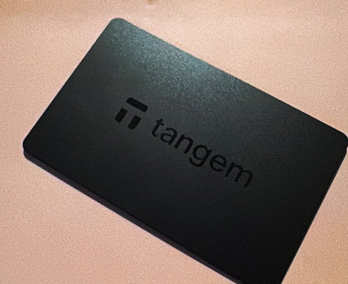 A dark green Tangem hardware cryptocurrency wallet card resting on a beige surface, displaying the Tangem logo and brand name in black text.