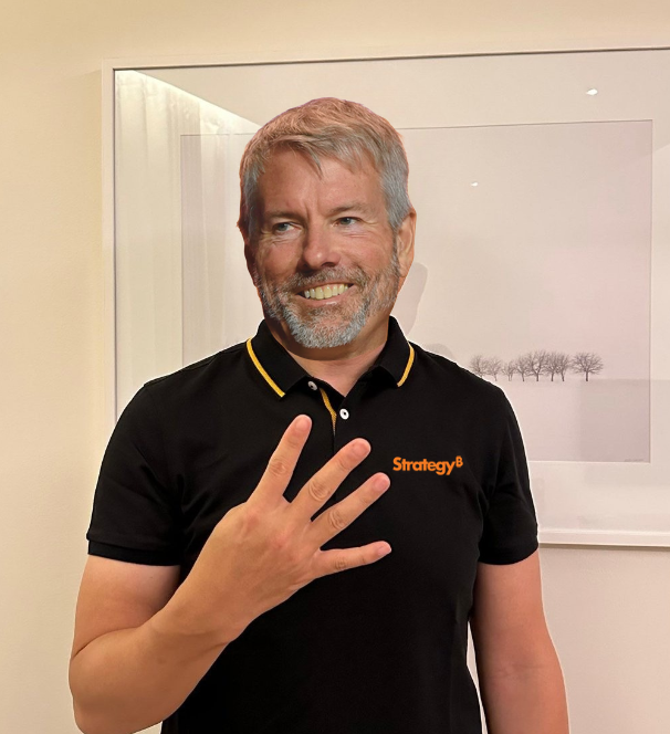 Michael Saylor, wearing a black 'Strategy' polo shirt with yellow trim, holds up four fingers in front of a framed landscape photograph.
