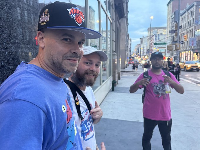 Two men pose for a selfie on a city sidewalk at dusk, with one wearing a New York Knicks cap and the other a white hat, while a third man in a pink shirt walks by in the background.