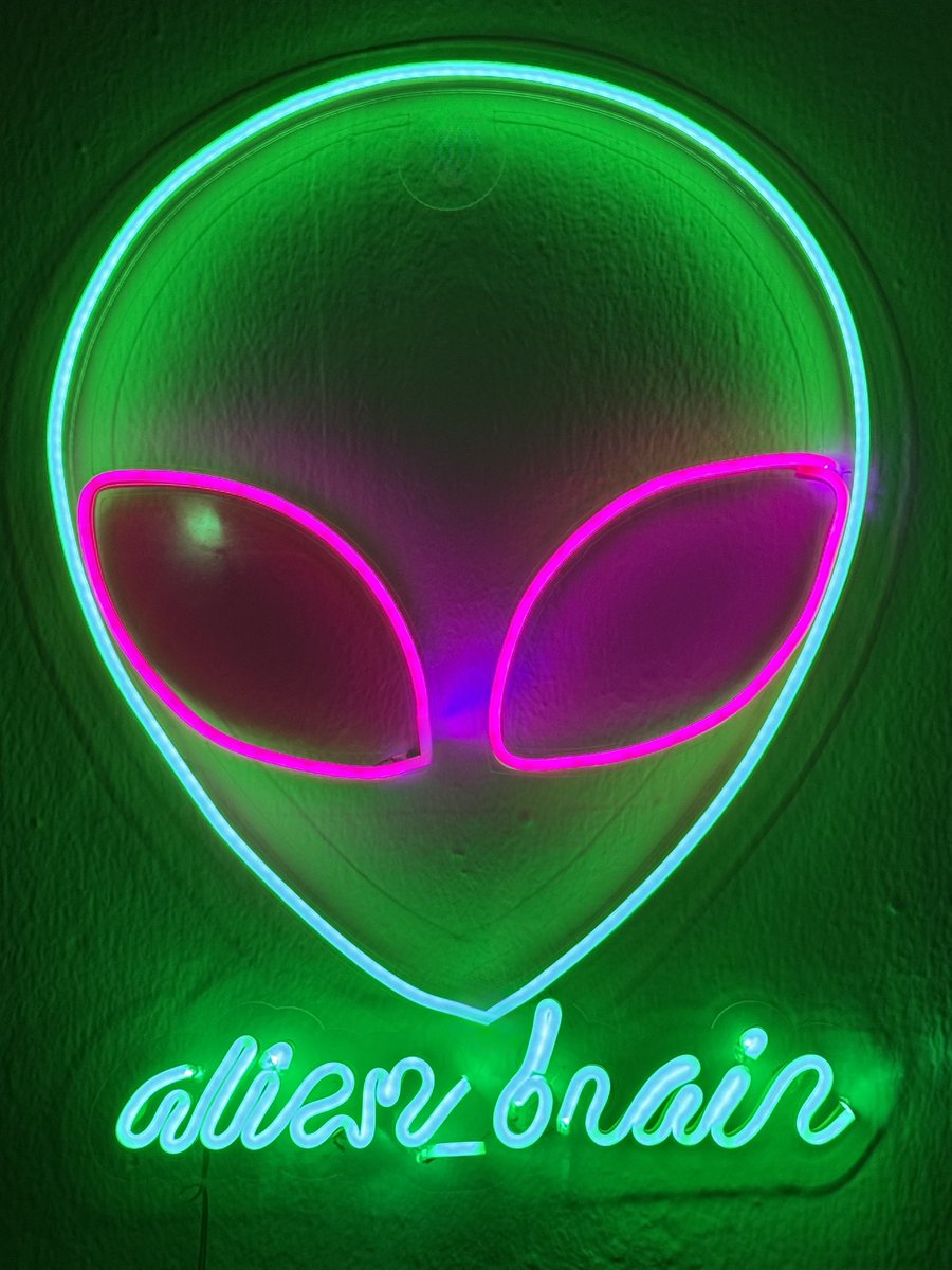 Neon sign in the shape of an alien head glowing green and pink against a green background, with reversed cursive text below that reads 'alien brain'.