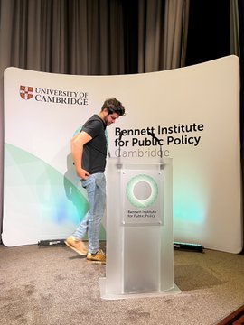 A man in a black shirt and jeans stands at a podium at the University of Cambridge's Bennett Institute for Public Policy, likely delivering a presentation, though the accompanying post humorously claims he has relocated to Monaco to recruit European artists for an NFT project called "VRNOUᑎS."