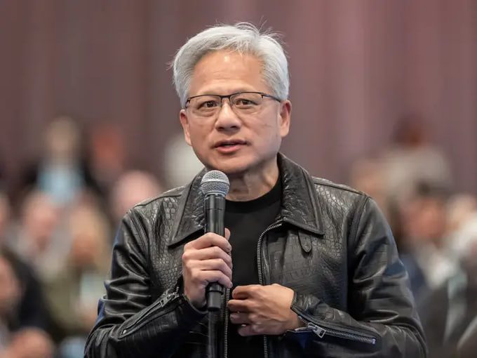 A man with silver hair and glasses, wearing a black leather jacket over a black shirt, speaks into a microphone while standing before a blurred audience.