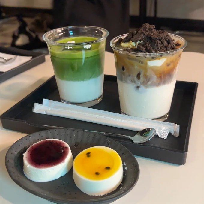A tray with two beverages—a matcha latte and an iced coffee topped with crushed Oreos—and a plate of two small cakes, one with red berry topping and one with passion fruit topping.