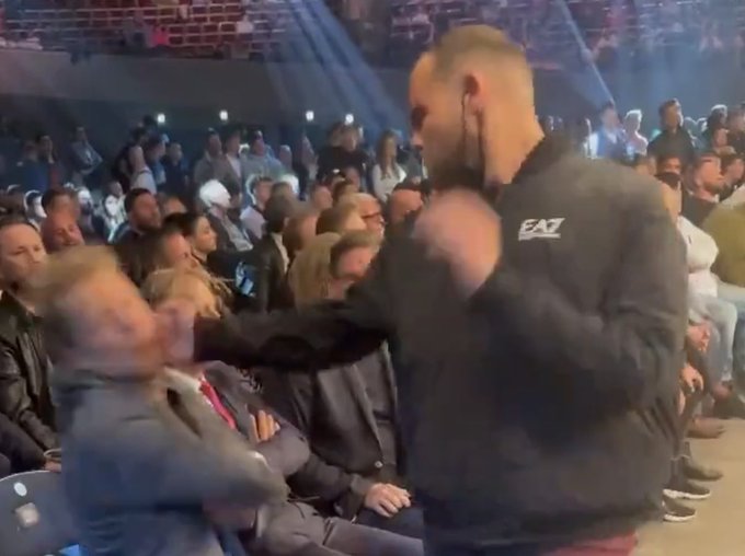 A crowded auditorium or stage area shows a man in a black “EA7” hoodie holding a microphone and facing seated audience members, with motion blur suggesting movement during the event.