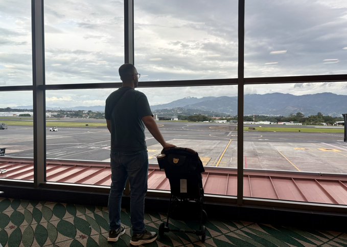 A man stands with luggage at an airport terminal window, looking out at the runway and mountains beyond.