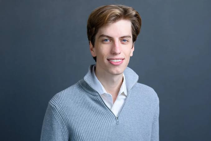 A young man with brown hair, wearing a gray zip-up sweater over a white collared shirt, smiles at the camera against a solid dark gray background.