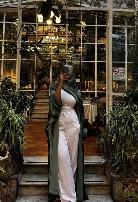 A woman wearing a long green hijab and white pants takes a mirror selfie with her phone in front of a glass-windowed venue decorated with hanging lights and potted plants.