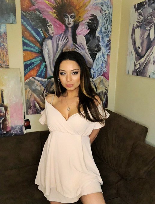 A young woman with long dark hair wearing a light pink off-shoulder dress, posing in front of colorful artwork and sitting on a brown couch.