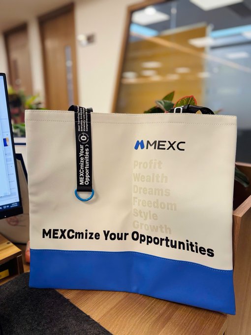 A cream and blue MEXC tote bag sits on a desk, featuring text including "MEXCmize Your Opportunities" and words like "Profit, Wealth, Dreams, Freedom, Style, Growth."