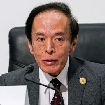 The image shows a man wearing a suit and tie sitting at a desk or table while speaking into a microphone. The background appears to be a white wall, which contrasts with the black leather chair he's sitting on. There are no other people visible in the photo. The main focus of the image is the man dressed in formal attire, suggesting that it might be related to an event or discussion involving financial matters.