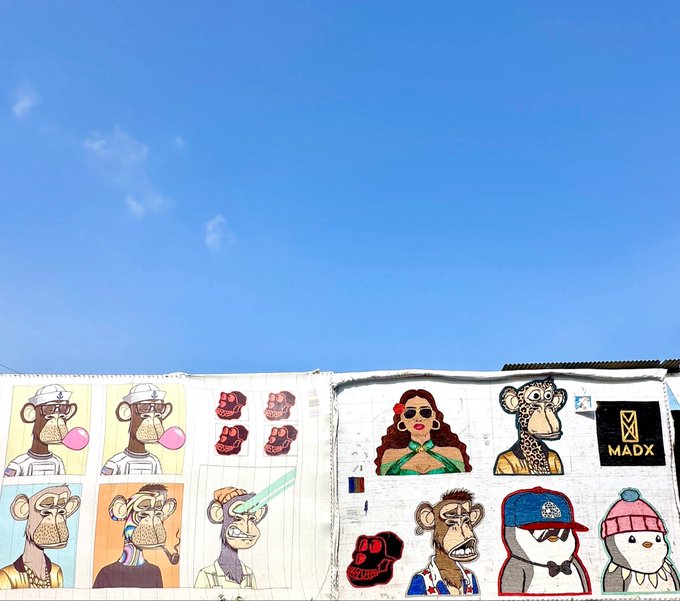 A vibrant outdoor mural displays a collection of stylized cartoon character portraits, including monkeys, ducks, and figures in various attire, along with the text "MADX" and a stylized "X" logo on a black square. The caption reflects the artist's meticulous process of creating these unique "masterpieces" one piece at a time, emphasizing their rarity and the patience of collectors awaiting the finished work.