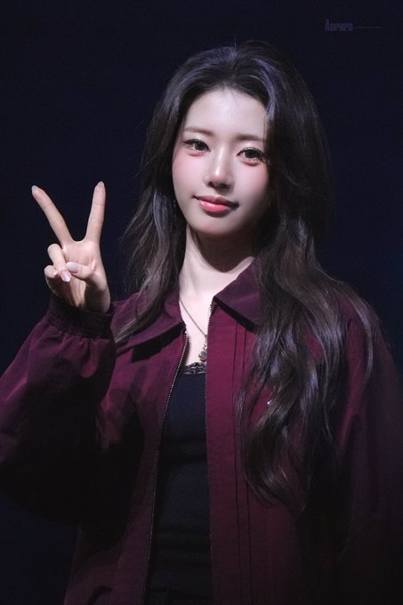 A young woman with long dark hair wearing a maroon jacket makes a peace sign, photographed against a dark background.