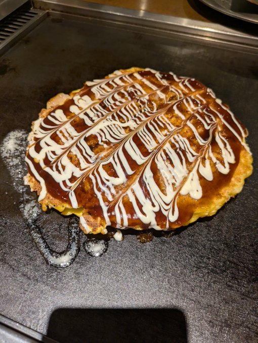 An okonomiyaki, a Japanese savory pancake, topped with a drizzle pattern of dark okonomiyaki sauce and white mayonnaise on a flat metal griddle.
