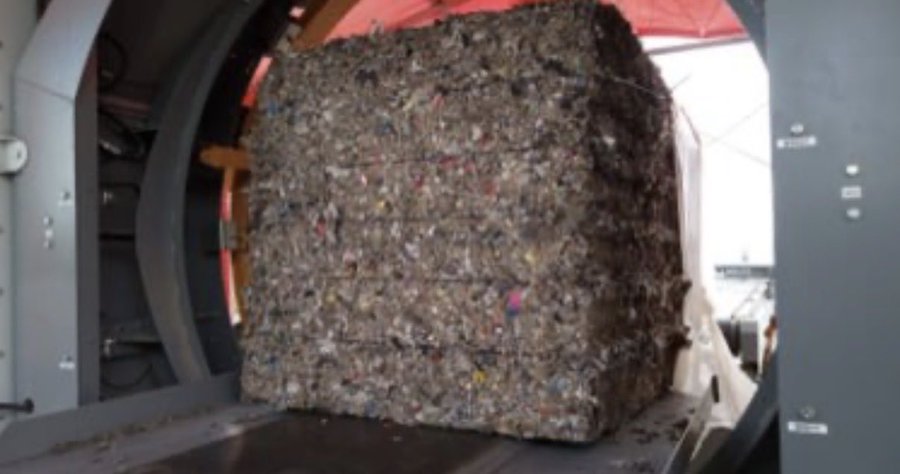 A massive compressed bale of recycled materials, including visible plastic bits, sitting inside an industrial processing machine.