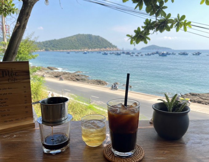 A Vietnamese coffee setup at Mộc Cafe featuring a phin filter, iced coffee, and tea, overlooking a bay dotted with boats and islands.