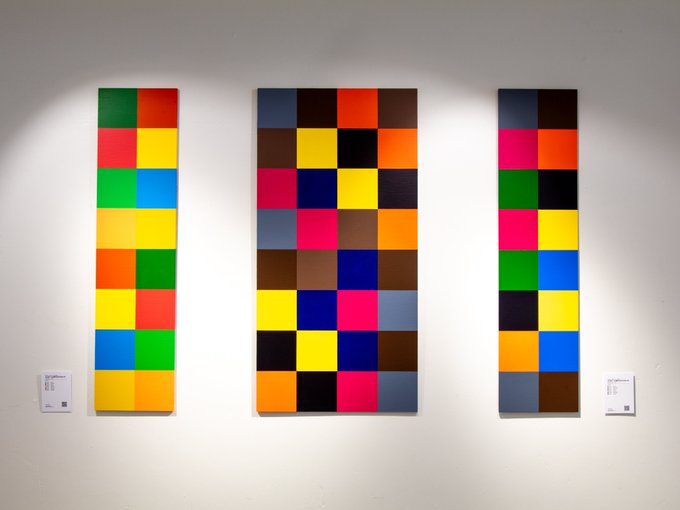 Three vertical abstract paintings displayed on a white gallery wall, each featuring grid-like compositions of brightly colored squares.
