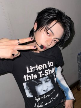 A young person with short dark hair, makeup, and black nail polish making a peace sign while wearing a black graphic t-shirt that reads 'Listen to this T-Shirt' featuring a printed image of a face.