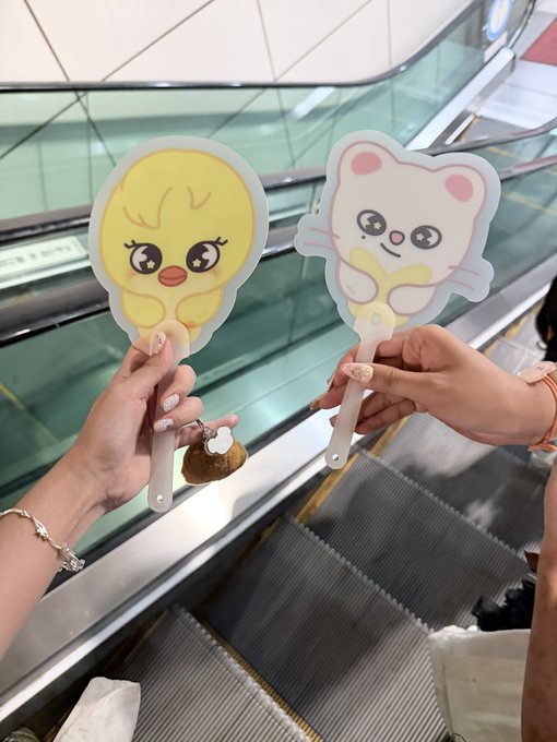 Two friends hold up cute character-themed fans (a yellow chick and a white bear with a pink bow) on an escalator, possibly trading merchandise like Leebit photocards. The playful hand fans suggest a fun shopping trip for K-pop collectibles.