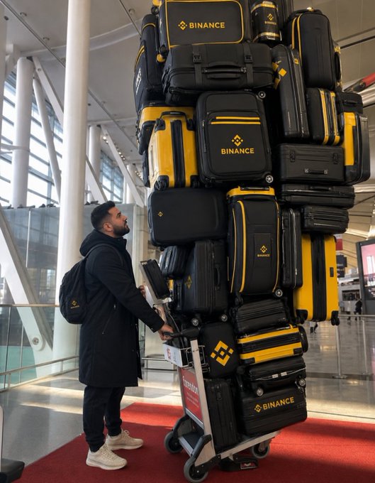 A bearded man in a black coat and backpack stands at an airport, gazing up at an enormous, towering stack of black and yellow Binance-branded suitcases precariously loaded onto a luggage cart.