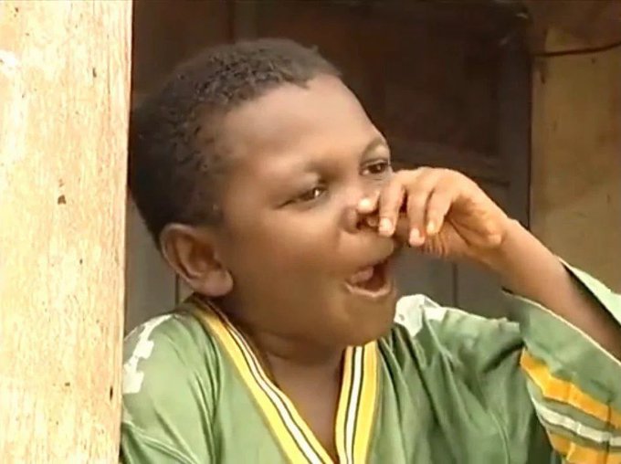 A young boy in a green shirt with yellow stripes laughs while covering his nose with his hand.