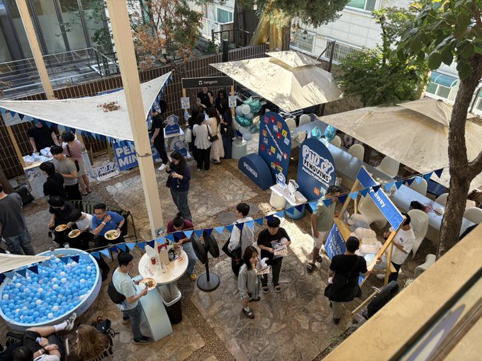 An overhead view shows an outdoor event space with Minions-themed decorations, a ball pit, people gathered around tables and food stalls, and a sign reading 'Longines'.
