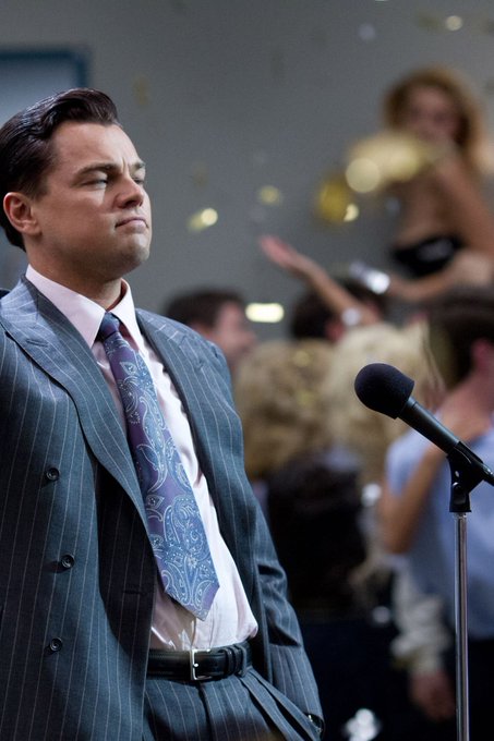 A sharply dressed man in a pinstripe suit and paisley tie stands solemnly at a microphone amid falling confetti, his expression reflecting the bittersweet weight of achievement, mirroring the post's cautionary message about Midas Market and perpetual trading: that what appears as user-driven success often masks a predetermined outcome, where the interface merely confirms what has already been decided, leaving the trader with the illusion of agency rather than genuine control.