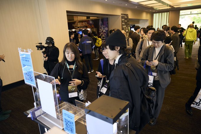 A crowded indoor event or conference with attendees wearing VIP badges, including a woman smiling while interacting at what appears to be a check-in or registration counter with QR codes.