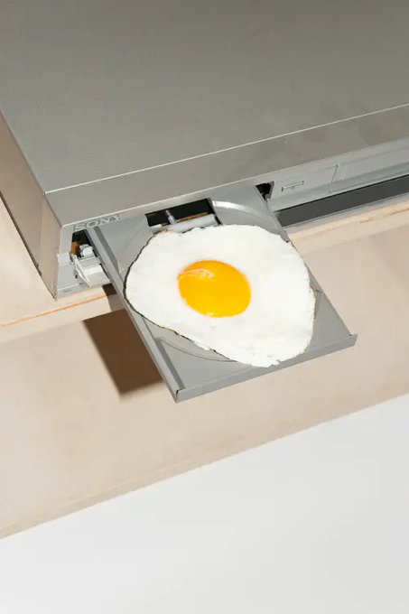 A realistic fried egg with a bright yellow yolk emerging from the disc tray of a silver CD/DVD drive mounted in a wooden surface.