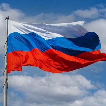 The Russian flag waving on a flagpole, displaying its white, blue, and red horizontal stripes against a blue sky with scattered clouds.