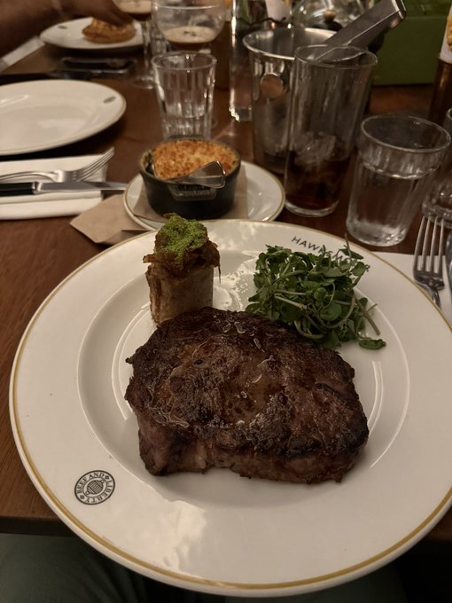 A perfectly seared ribeye steak plated with bone marrow topped with herb gremolata and fresh microgreens, served at a steakhouse (with a side dish visible in the background). The image embodies the "eat steak" and "build muscle" lifestyle pillars, presenting a premium, high-protein meal as an aspirational reward in the creator's daily ritual.