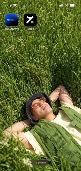 A phone home screen showing 9:00 with apps TOKEN2049 and Hotcoin, featuring a wallpaper of a person in a green vest and orange sunglasses lying in a grassy field.