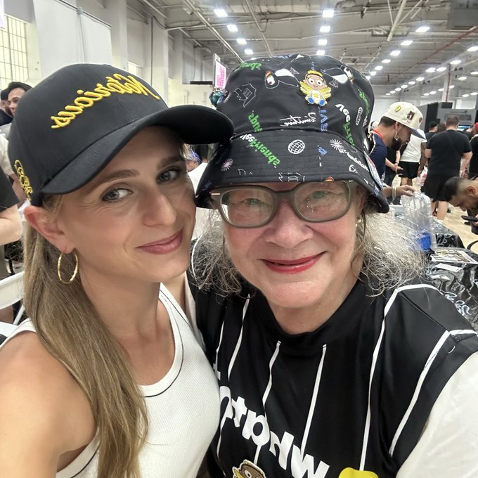 A selfie of two women, one wearing a black cap with yellow text and the other wearing a patterned bucket hat and glasses, posing together at what appears to be a convention.
