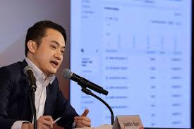 The image shows a man sitting at a podium with a microphone, giving a speech or presentation. The setting appears to be an event where he is addressing an audience. The focus of his speech seems to be stablecoins, as they are being discussed in relation to their importance and growth within the digital asset industry.