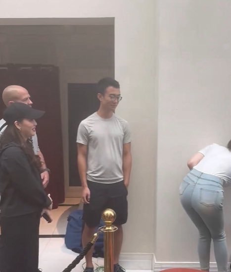 A young man with glasses in a gray t-shirt and black shorts stands in a hallway near a woman in a black cap and another person bending over, with the post humorously suggesting he's cashing out his $HYPE crypto holdings to spoil someone with luxury purchases.