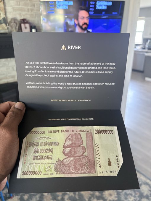 A hand holds an open black presentation folder from "RIVER" containing a hyperinflated Zimbabwean banknote (200 million dollars) with explanatory text comparing traditional currency inflation to Bitcoin's fixed supply model.