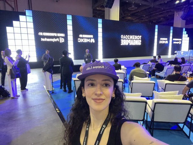 A woman wearing a purple 'Kelpdao Lab' cap takes a selfie at what appears to be a WebX Singapore event, with attendees seated in the background and large screens displaying 'SINGAPORE TOKEN2049' and 'WebX'.