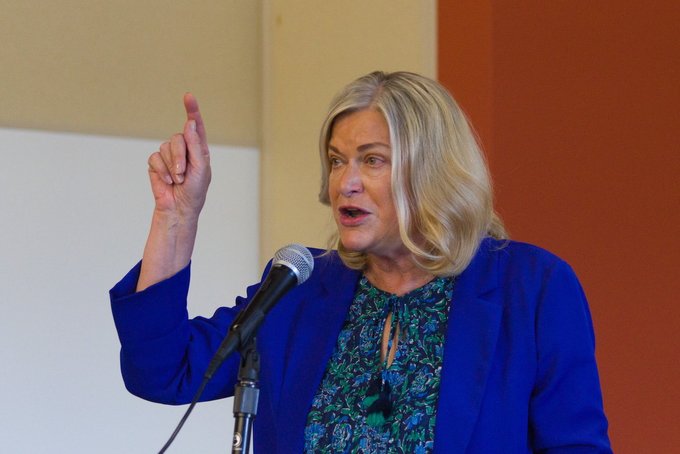 A blonde woman in a blue blazer speaks passionately into a microphone with her right index finger raised, appearing to be U.S. Senator Cynthia Lummis as she discusses the CLARITY Act and its aim to nationalize Wyoming's crypto-friendly regulatory framework for digital assets.