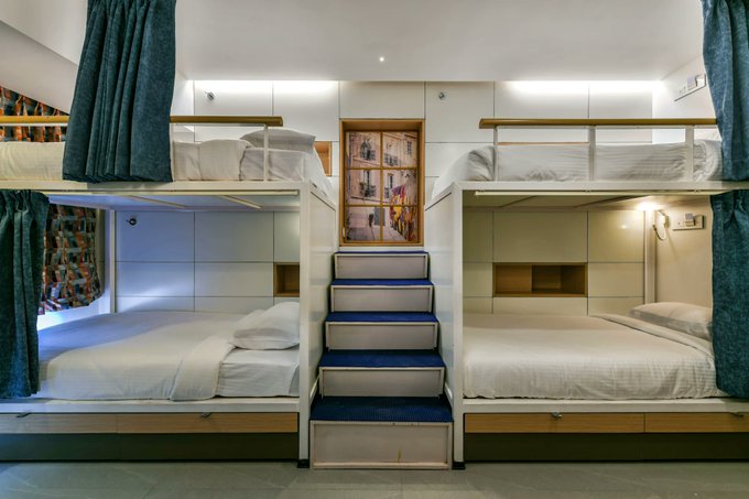 A modern dormitory room featuring four bunk beds with privacy curtains, white linens, and a central staircase, providing shared accommodation for the Dev3pack Residency attendees in Mumbai during Devcon India.