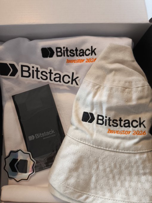 A photo showing merchandise in a box, including a t-shirt, a black item in packaging, a sticker, and a bucket hat, all branded with 'Bitstack Investor 2026'.