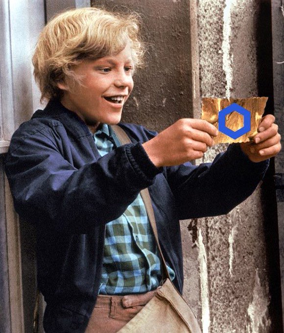 Still from a film showing a blonde-haired boy in a blue jacket and plaid shirt excitedly holding a golden ticket or card with a blue hexagonal logo.
