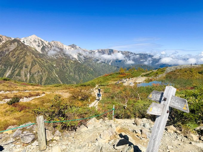 A scenic mountain landscape with rugged peaks, low clouds, and hikers walking along a rocky trail beside a wooden sign, with autumn-colored vegetation in the foreground.