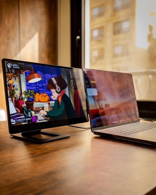 A portable monitor sits on a wooden desk next to a laptop, displaying an animated scene of a person working, with a window overlooking a city in the background.