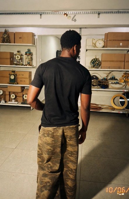 A man in a black t-shirt and camo pants stands with his back to the camera, browsing what appears to be a clock and décor shop with shelves of timepieces visible. The post "Gm CT lets WIN" suggests a casual morning greeting, fitting the relaxed vibe of someone perusing a store early in the day, though no specific text or numbers are visible in the image itself aside from a date stamp "10/06" in the corner.