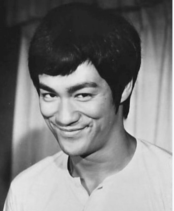The black-and-white photo shows a smiling young man with dark hair wearing a white shirt, capturing a candid and cheerful moment. The image reflects the inspirational spirit of the accompanying quote, embodying Bruce Lee's resilient and positive mindset that refuses to accept defeat.