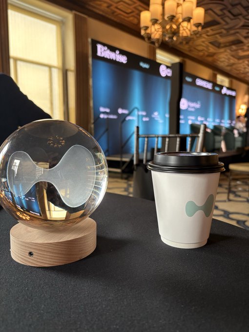 A crystal ball etched with a bowtie logo sits beside a branded paper coffee cup on a table, with an event backdrop displaying "Bitvavo" and "Hyperliquid" visible in the background, suggesting the scene captures a branded moment at a crypto industry conference.