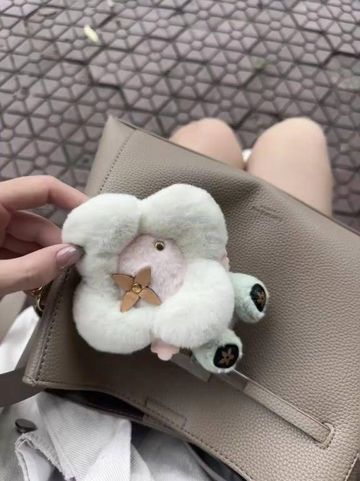 The image features a person holding up a small stuffed animal, which appears to be a cute flower-shaped plush toy made of white fur. The person's hand is positioned over the toy, showcasing its details. The background of the image is blurred, drawing attention to the main subject - the stuffed animal and the person holding it.