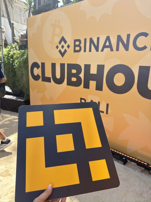 A hand holds up a Binance logo sign in front of a bright orange "BINANCE CLUBHOUSE BALI" event banner, capturing the vibrant atmosphere of the #BinanceClubhouse at Coinfest 2026.