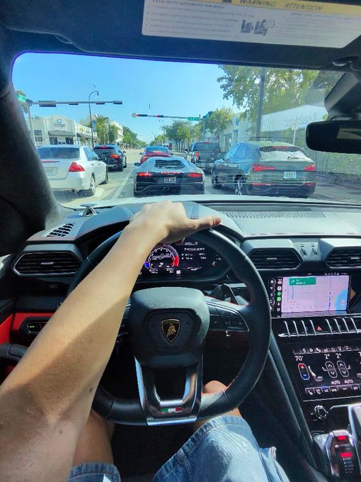 First-person view from inside a Lamborghini, showing a hand on the steering wheel with the iconic bull logo, driving down a sunny street with other cars and a navigation screen visible on the dashboard.