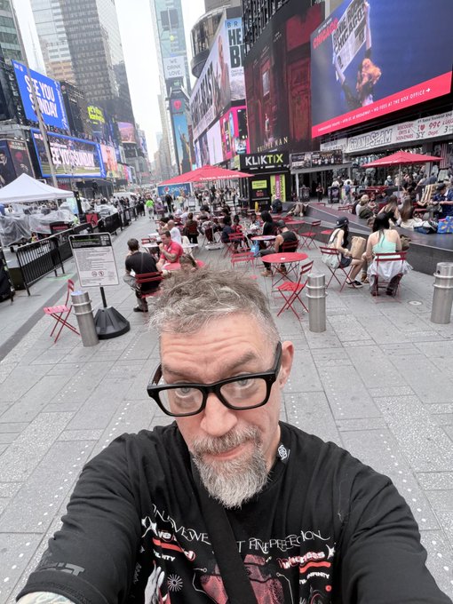 A selfie of a man with glasses and a goatee in Times Square, New York, with outdoor seating areas and bright billboards visible in the background.