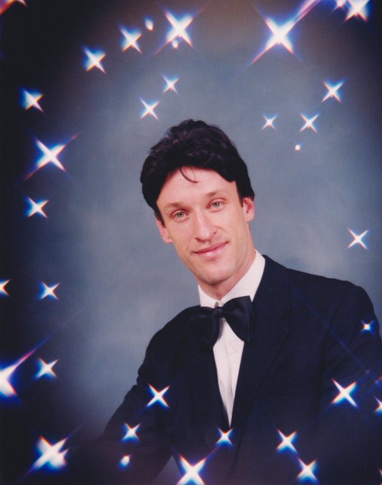 A man in a black tuxedo and bow tie smiles against a backdrop of bright, star-shaped light flares.